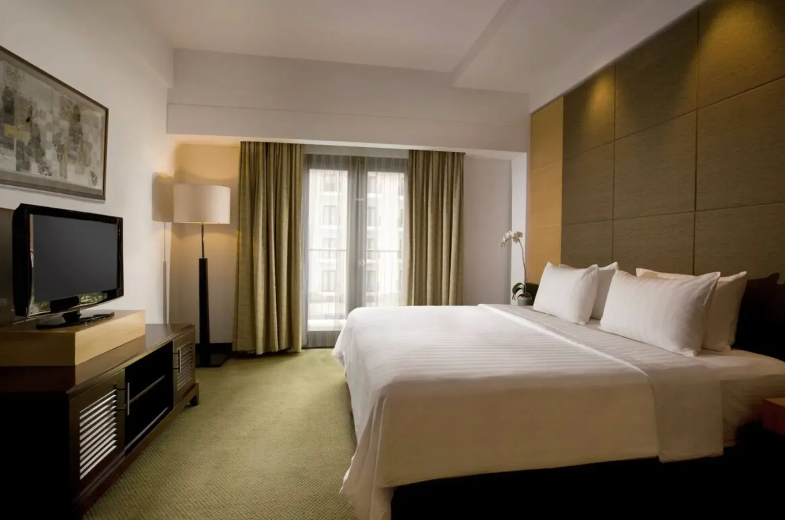 Hotel guest room with finished painted drywall, a padded fabric accent wall behind the bed, a curtained balcony door, a wall-mounted television, and neutral furnishings