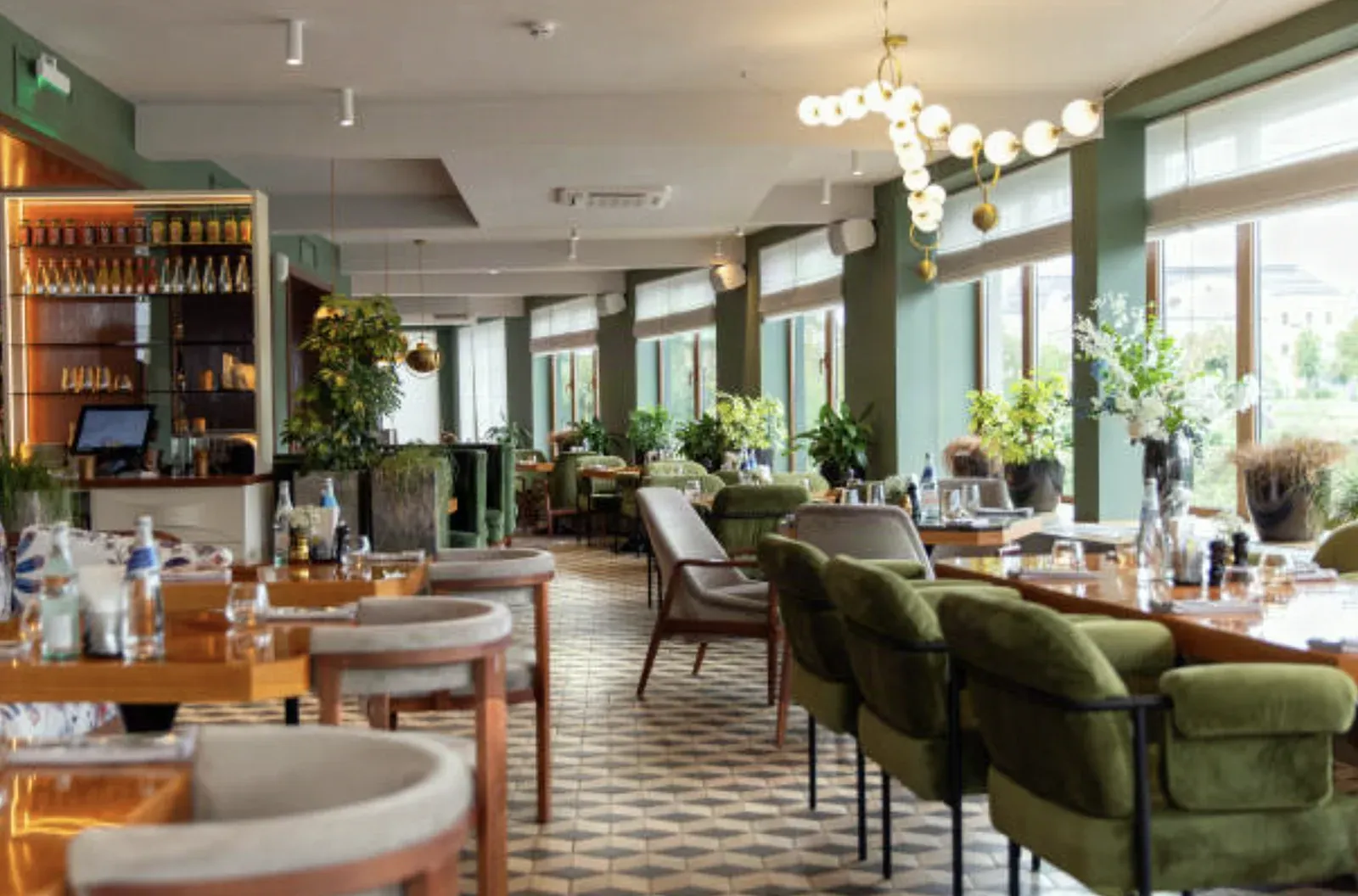 Restaurant dining room with a stepped drywall ceiling soffit, sage-green window piers, a glass-globe chandelier, and green velvet chairs on patterned tile
